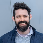 Smiling man with dark hair, beard, and mustache wearing a gray sweater and dark jacket.