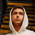 Young man with curly hair wearing a white hoodie, looking slightly upward with a serious expression, sitting on wooden steps.