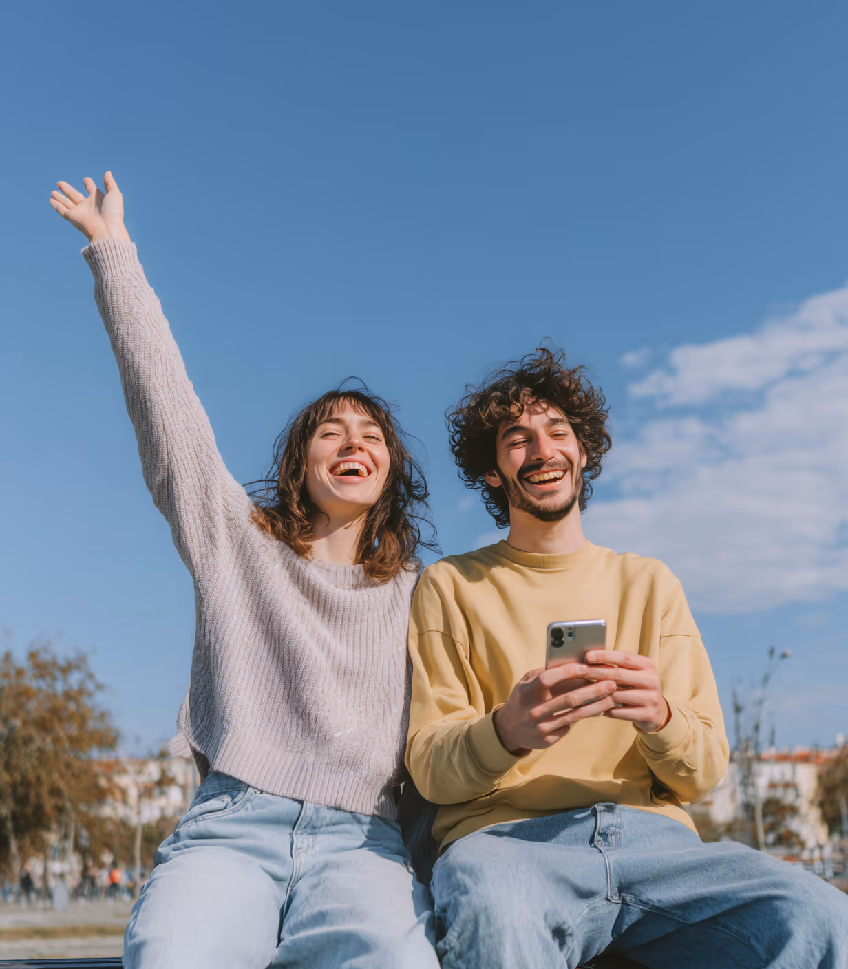 Happy young woman raising her arm and smiling next to a young man holding a smartphone outdoors under a blue sky.