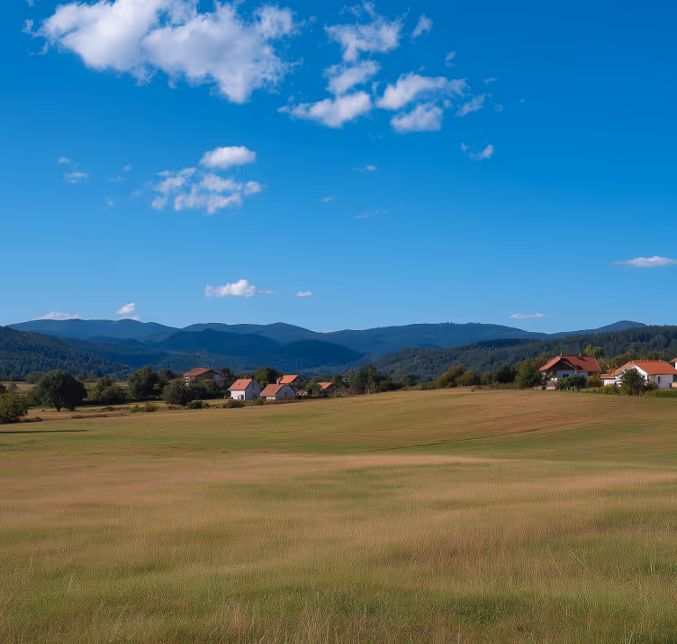 Rural landscape with green fields, scattered houses with red roofs, and mountains under a blue sky with clouds.