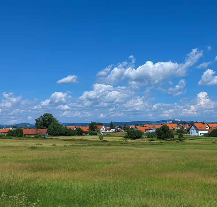 Rural landscape with green fields, scattered houses with red roofs, and mountains under a blue sky with clouds.