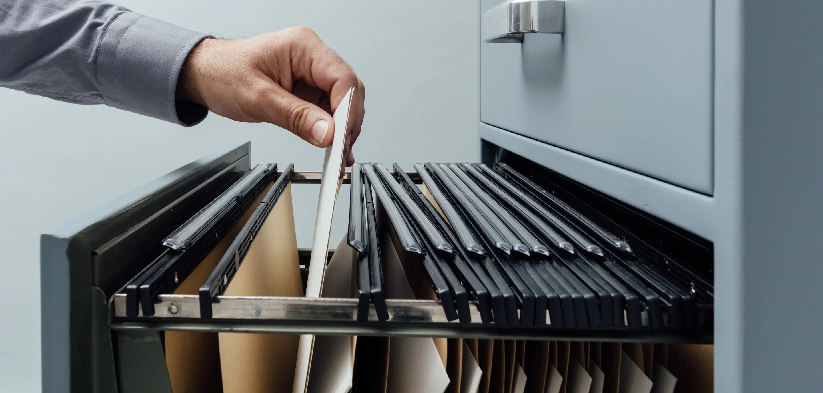 Hand pulling a file folder from an open gray filing cabinet drawer filled with hanging folders.