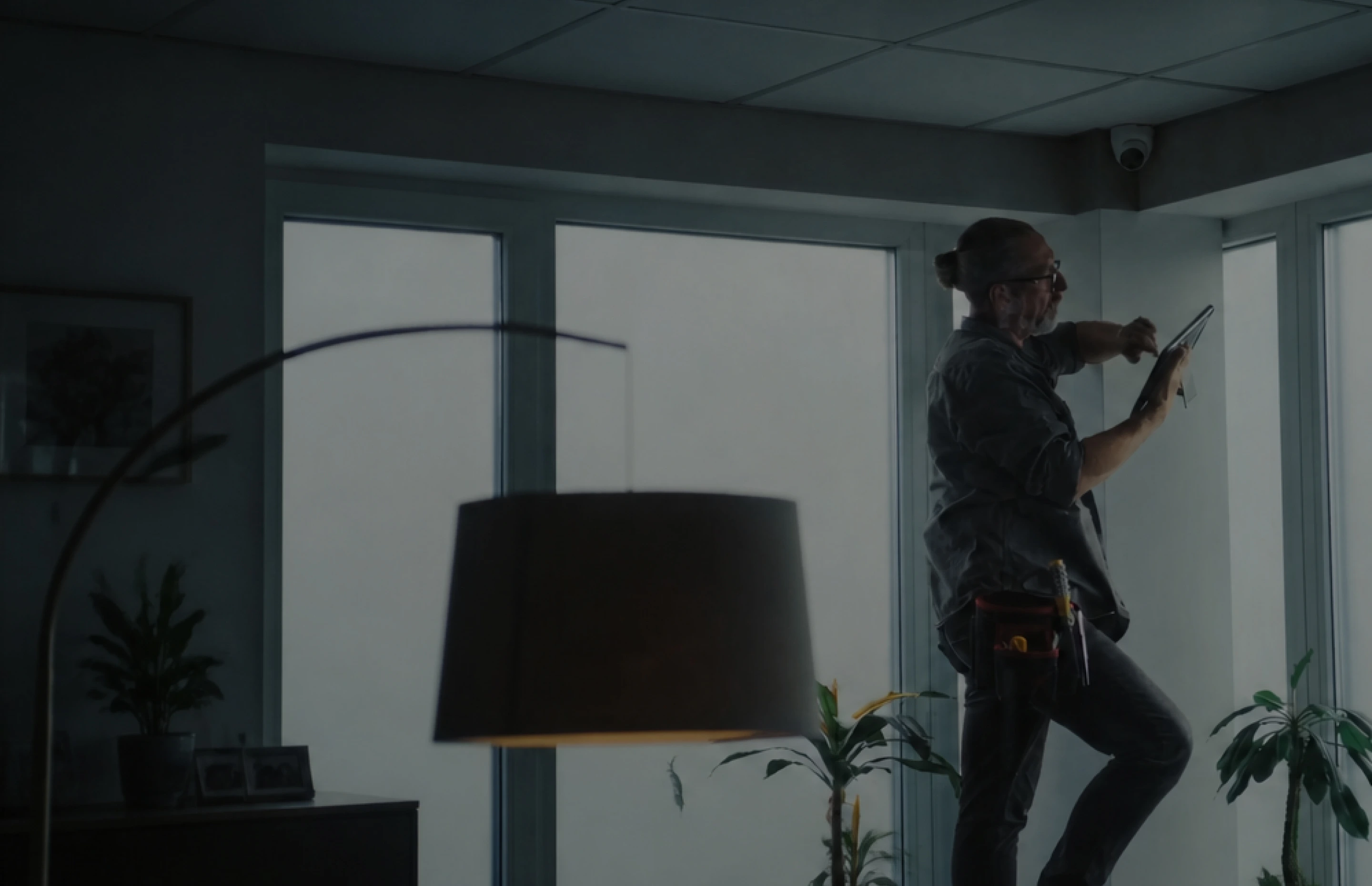 Man with tools inspecting a wall-mounted device in a room with large windows and plants.