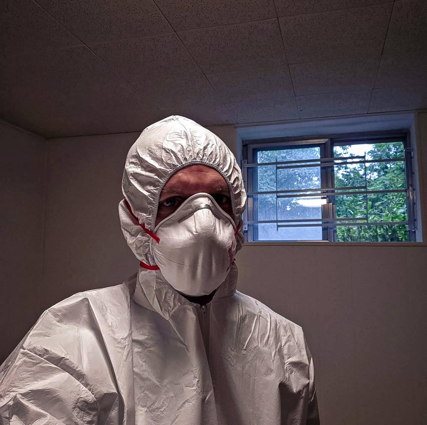 Person wearing a white protective suit with a hood and a respirator mask indoors near a barred window.