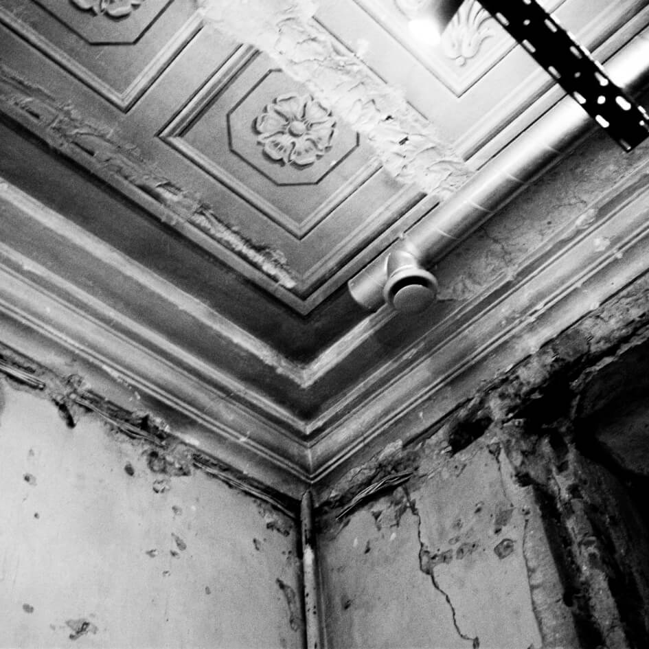 Corner of an old room with peeling paint, cracked walls, decorative ceiling panels, and exposed pipes.