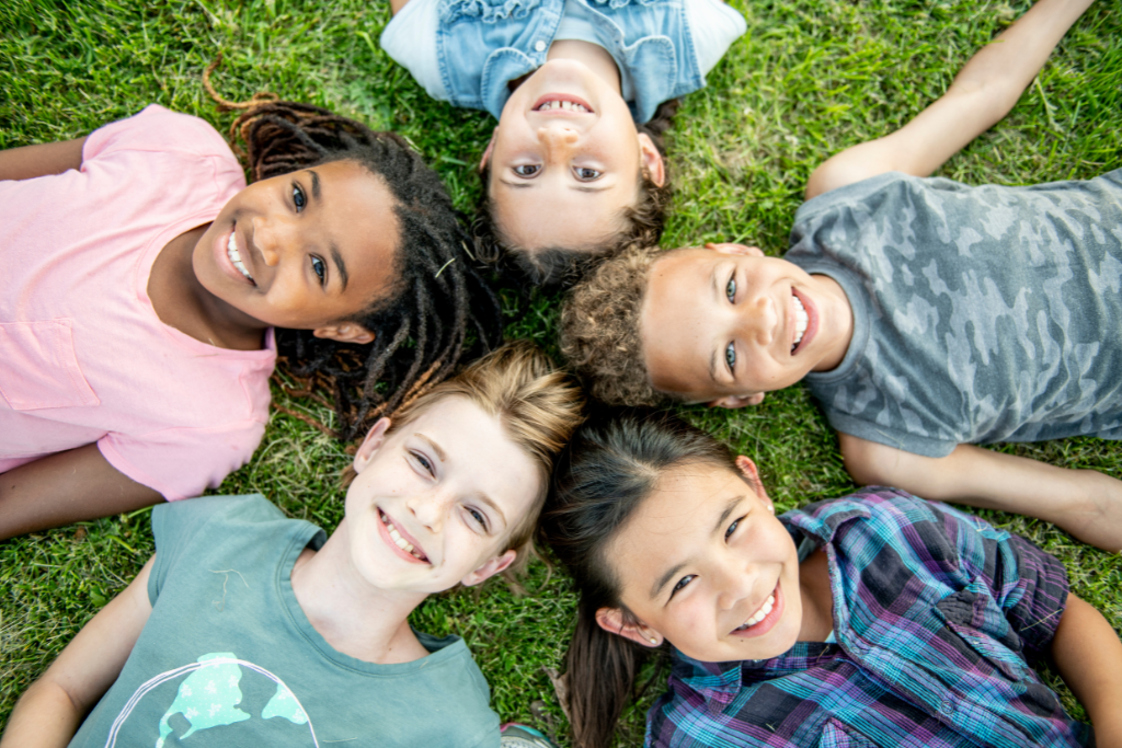 Group of diverse children lying on grass smiling together, representing Regulation First Parenting™ and the benefits of nervous system regulation for kids.