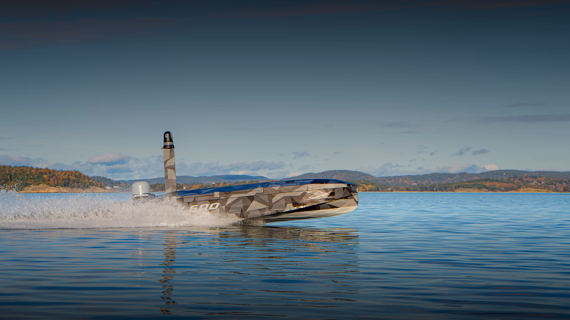 Camouflaged unmanned surface vehicle speeding on a calm lake with forested hills in the background.
