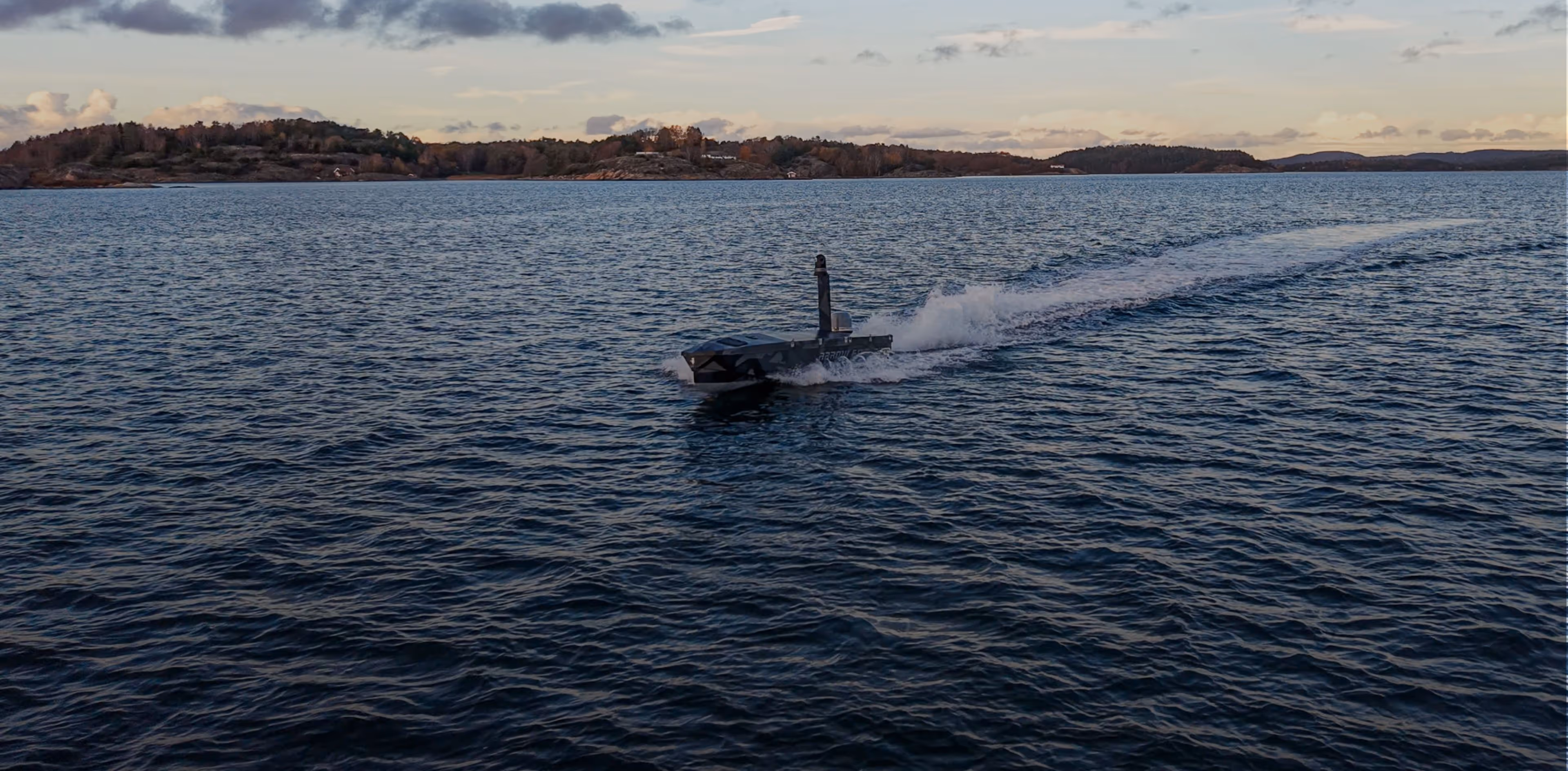 Autonomous surface vessel moving through calm open water near a forested shoreline under a cloudy sky.