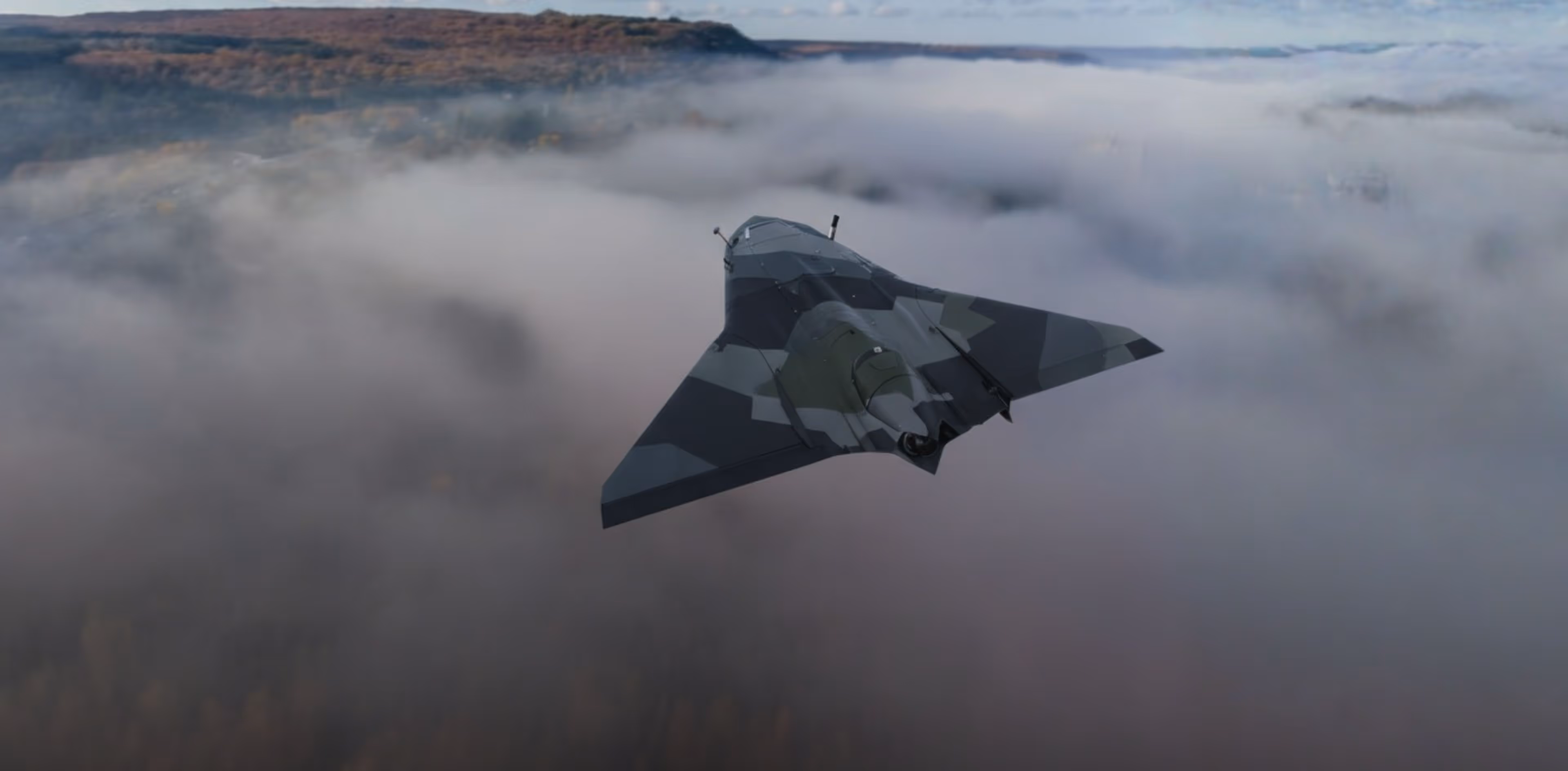 Camouflaged military drone flying above fog with hills in the background.
