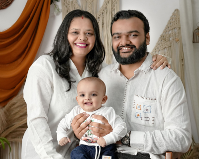 Smiling couple poses with their baby, all dressed in white clothing, indoors with warm decor.