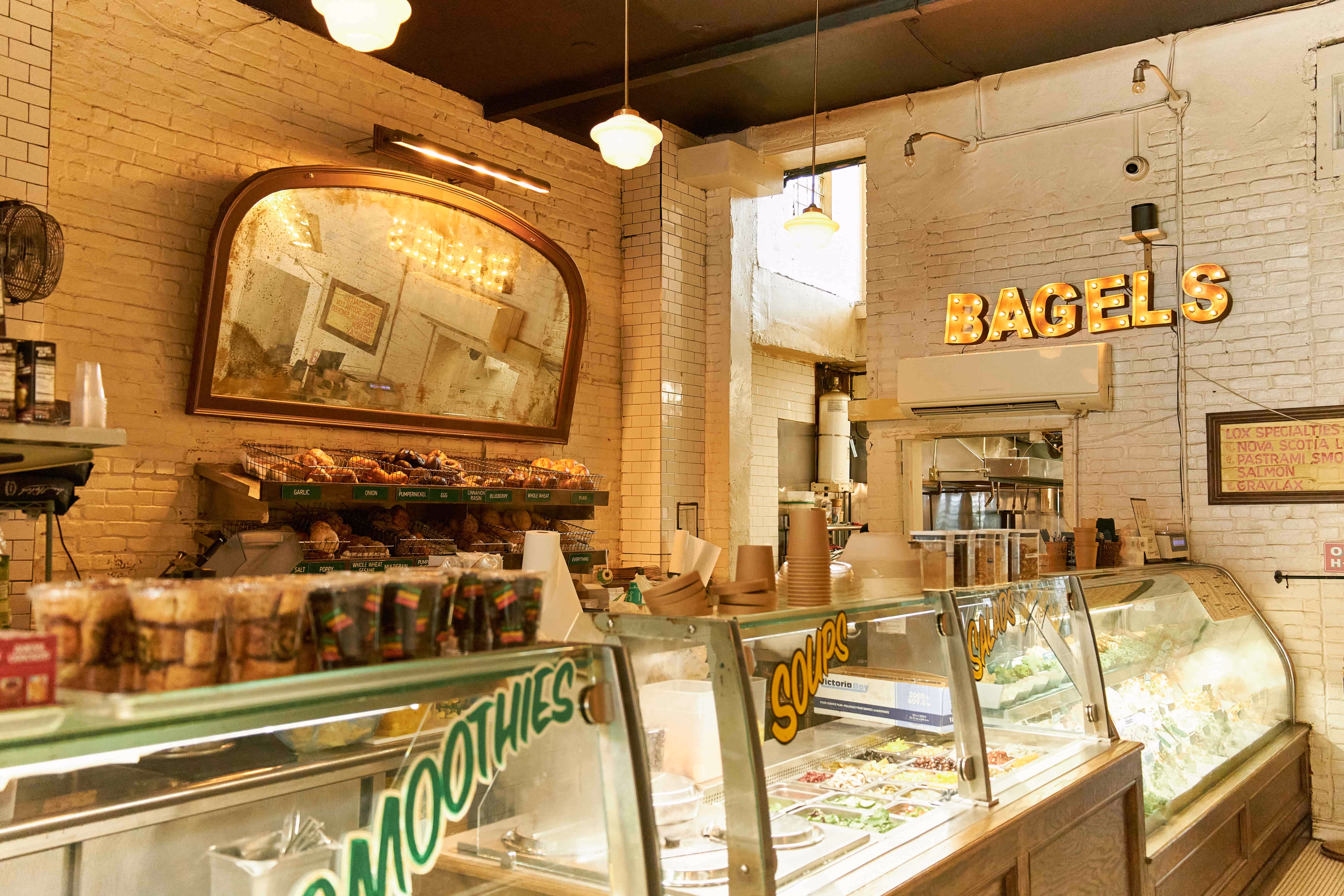 Interior of a bakery with a glass display counter containing salad and smoothie ingredients, a wooden shelf with various bagels, and a lit sign reading 'BAGELS' on a white brick wall.