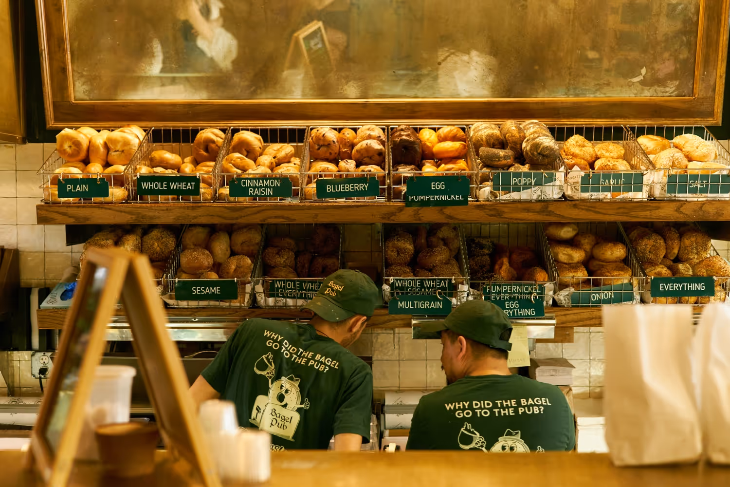 Bagel shop counter with assorted bagels and staff in green uniforms.