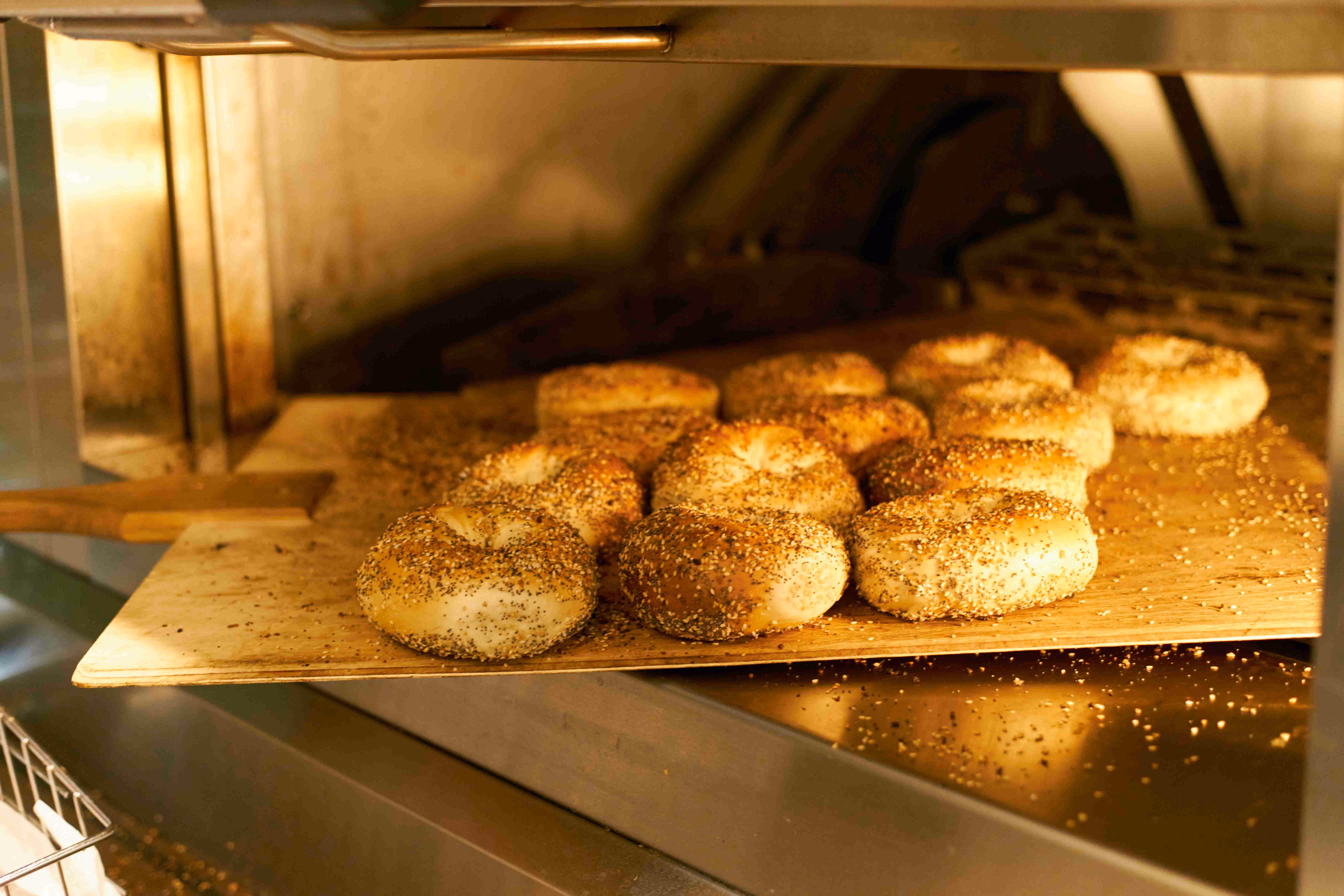 Several sesame and poppy seed bagels baking in a commercial oven on a wooden peel.