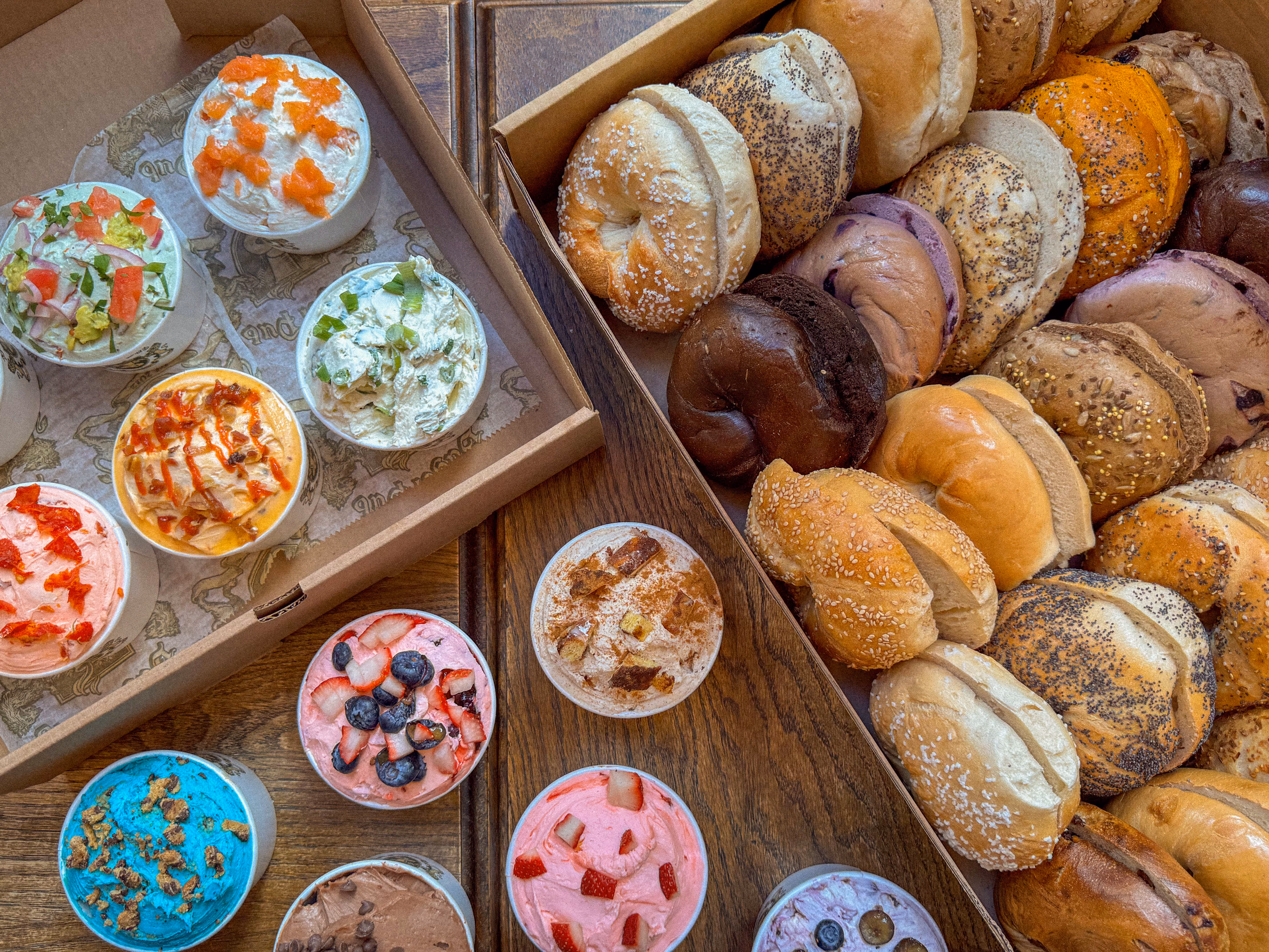 Variety of bagels in a box next to cups filled with colorful cream cheese spreads topped with ingredients like fruit, herbs, and spices.