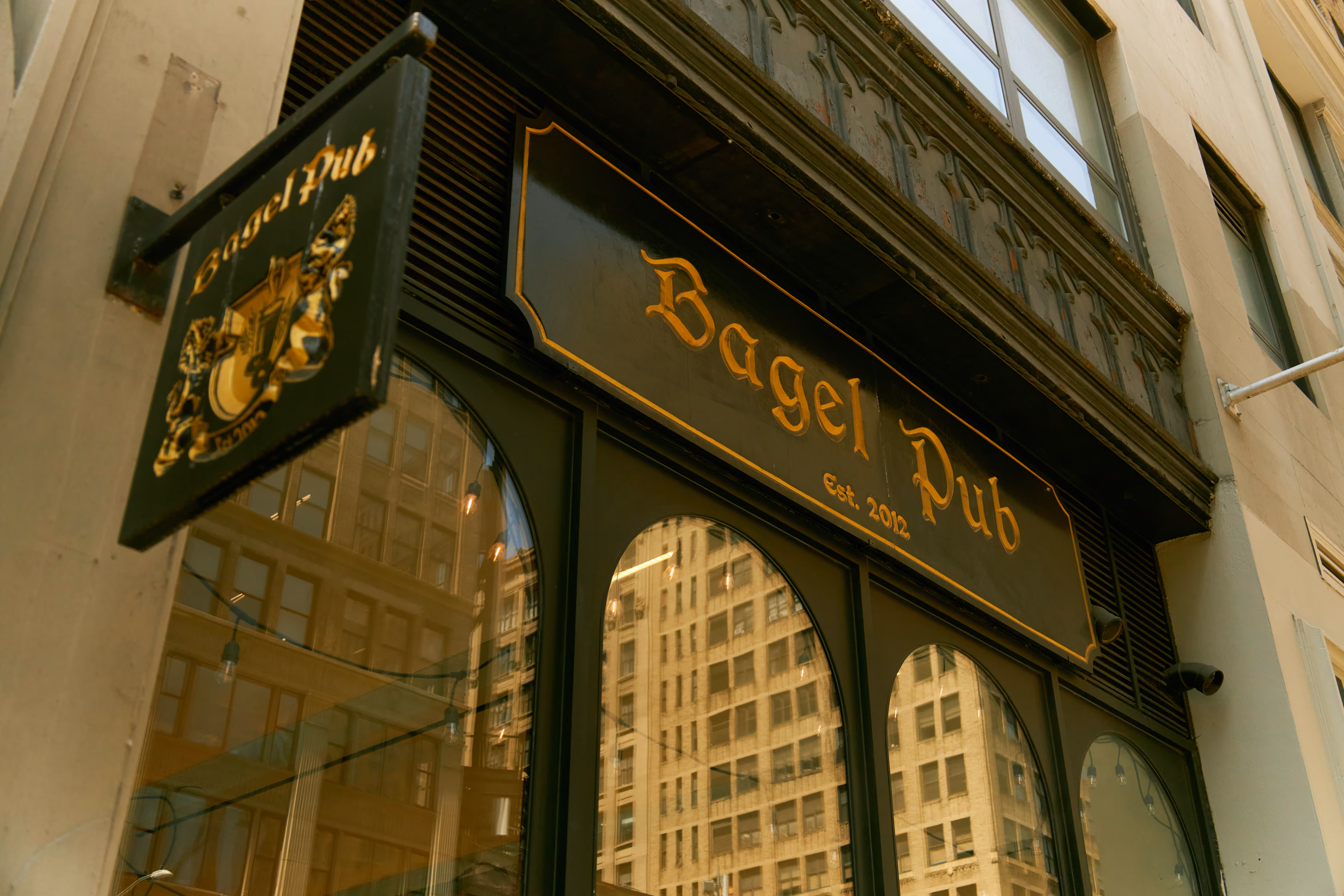 Black and gold sign reading 'Bagel Pub est. 2012' above arched windows reflecting nearby New York buildings.