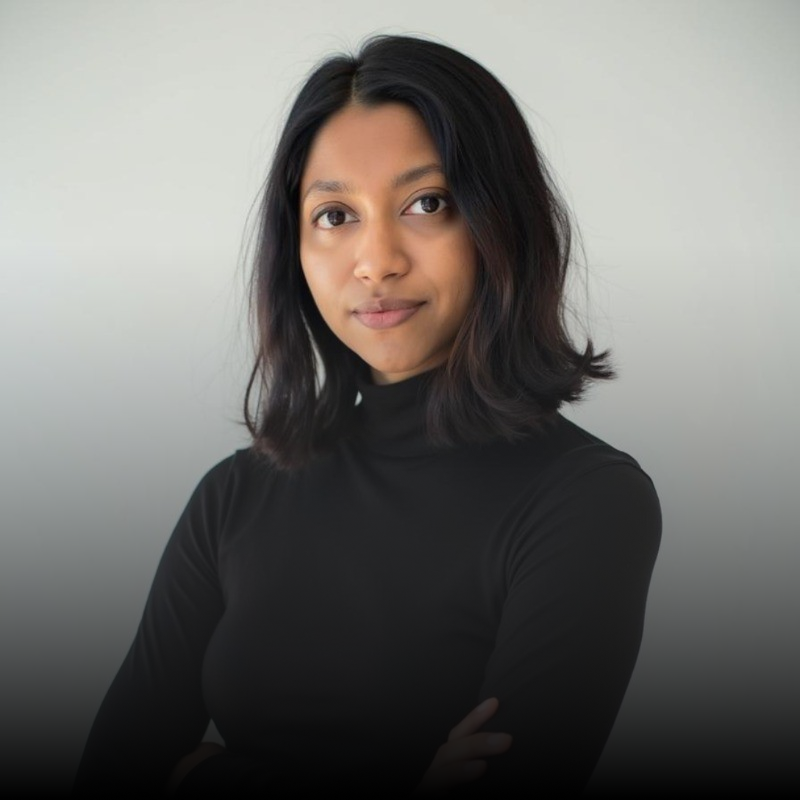 Portrait of a confident woman with shoulder-length black hair wearing a black turtleneck against a light gray background.