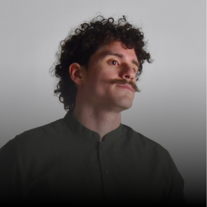Portrait of a man with curly hair and a thick mustache wearing a dark buttoned shirt, looking slightly upward.