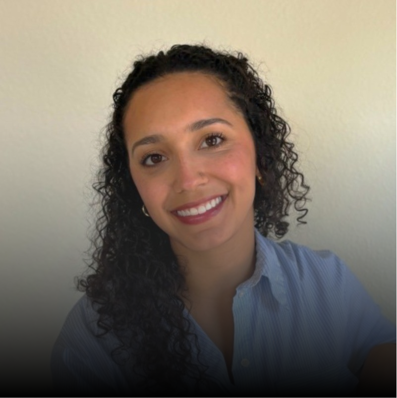 Portrait of a smiling woman with curly dark hair wearing a light blue shirt against a plain light-colored background.