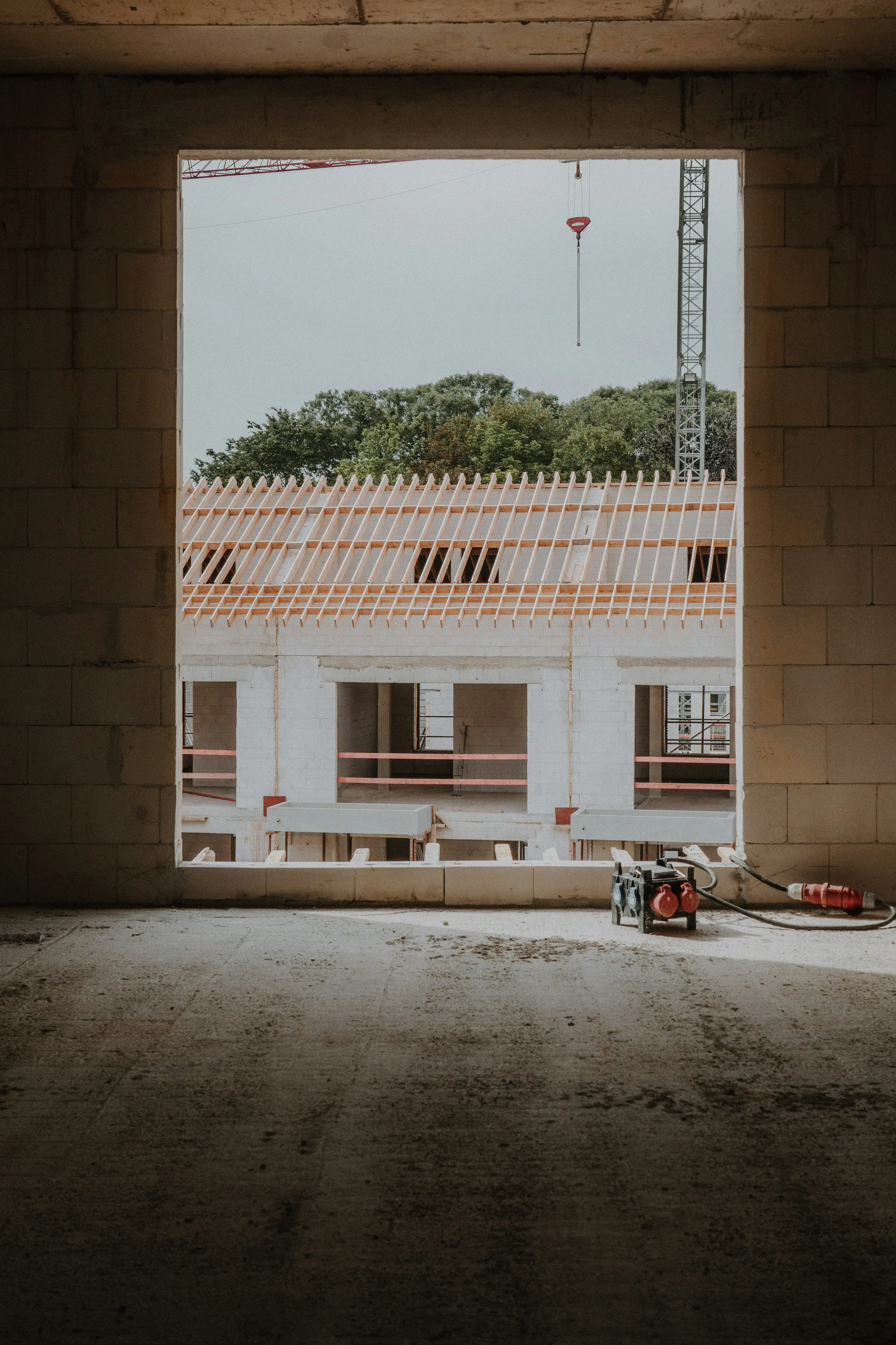 Kilátás egy befejezetlen ablakkereten keresztül egy építés alatt álló épületből, a háttérben daruval és fákkal.