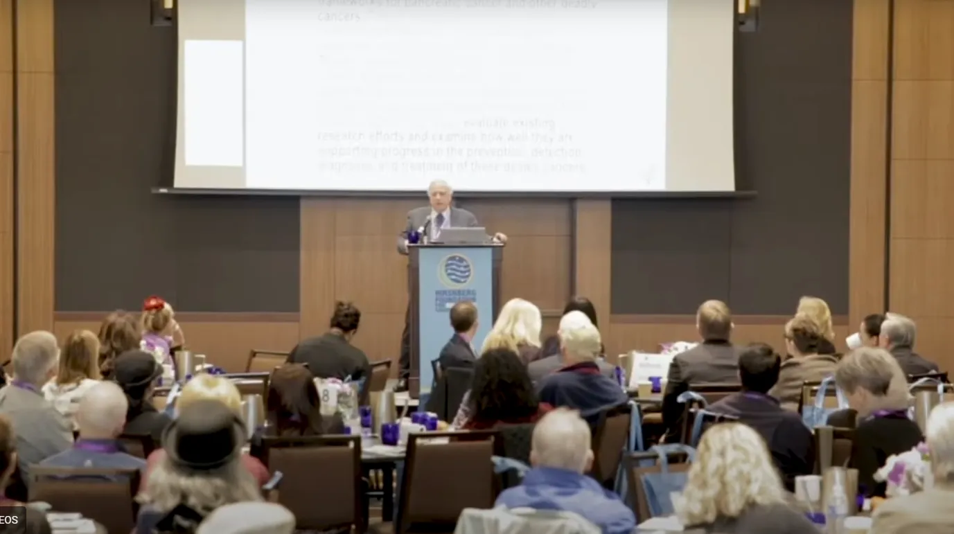 Speaker at podium addressing an audience seated at round tables in a conference room.