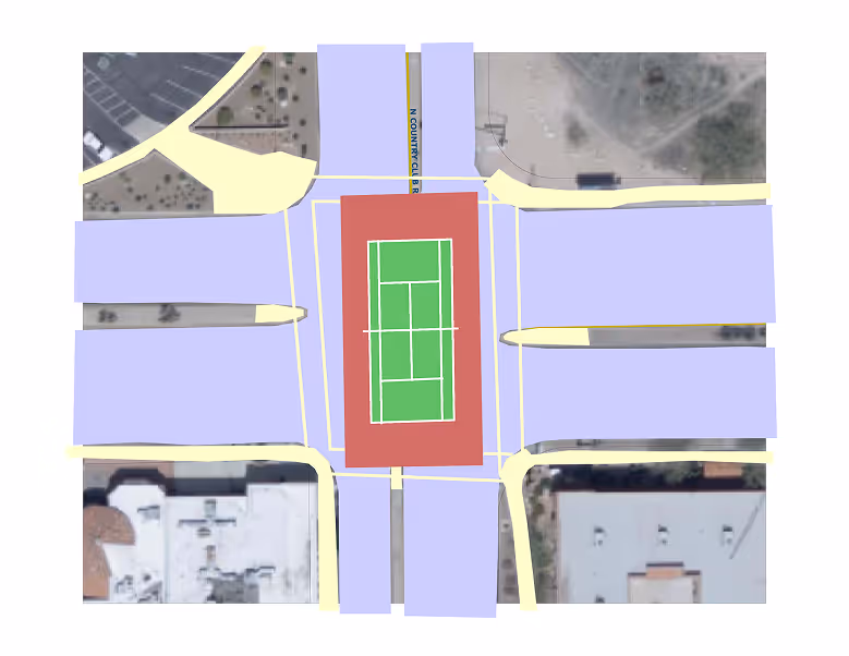 Top-down map view of an urban intersection with a central tennis court marked in green surrounded by red and blue areas representing roads and buildings.