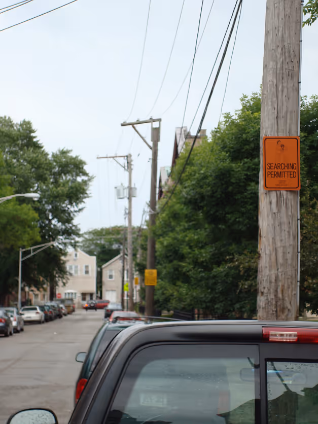 Residential street lined with parked cars, utility poles, and a sign on a pole reading 'SEARCHING PERMITTED'.