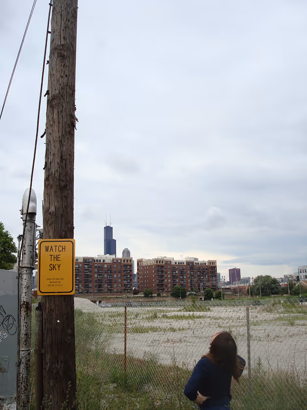Person looking up at a cloudy sky near a utility pole with a sign reading 'WATCH THE SKY' and a city skyline in the background.
