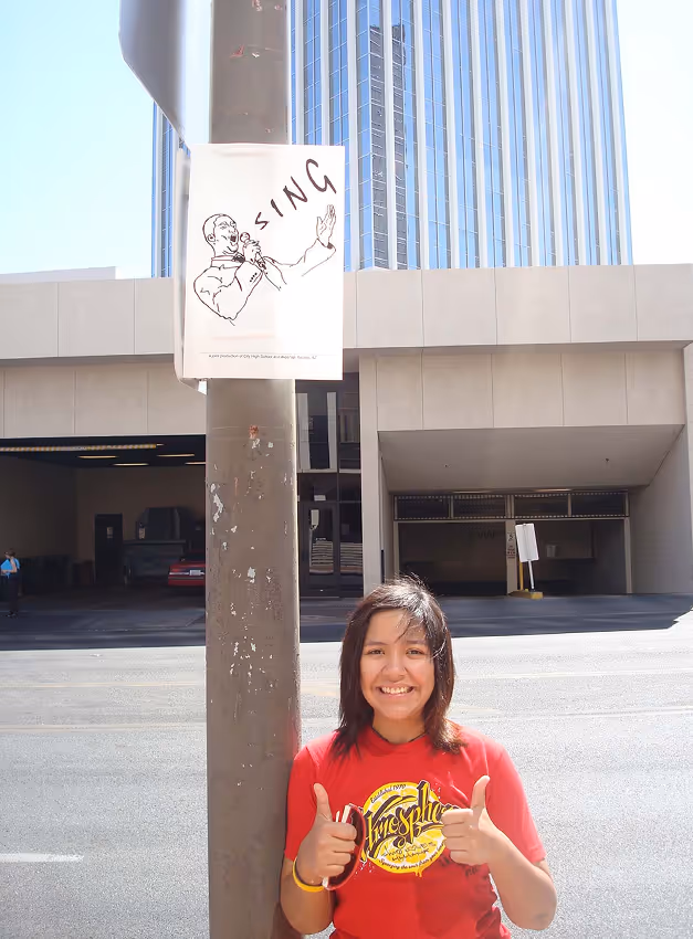 Young woman in a red shirt giving two thumbs up standing next to a pole with a sign that reads 'SING' and a drawing of a person singing, with a tall building in the background.