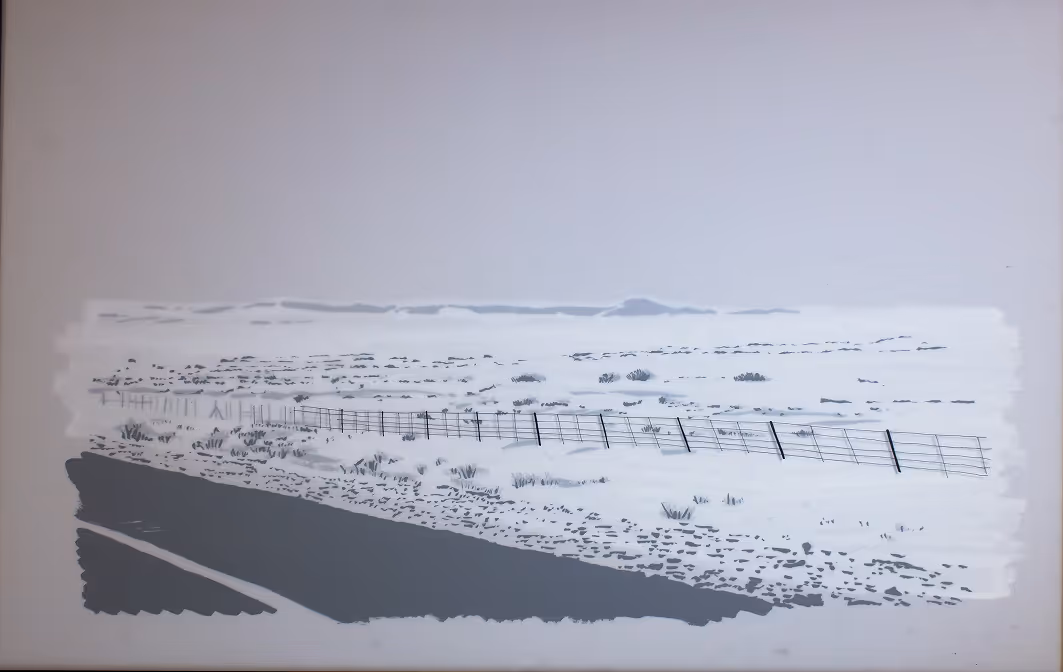 Empty road along a snowy landscape with sparse shrubs and a wire fence extending into the distance.