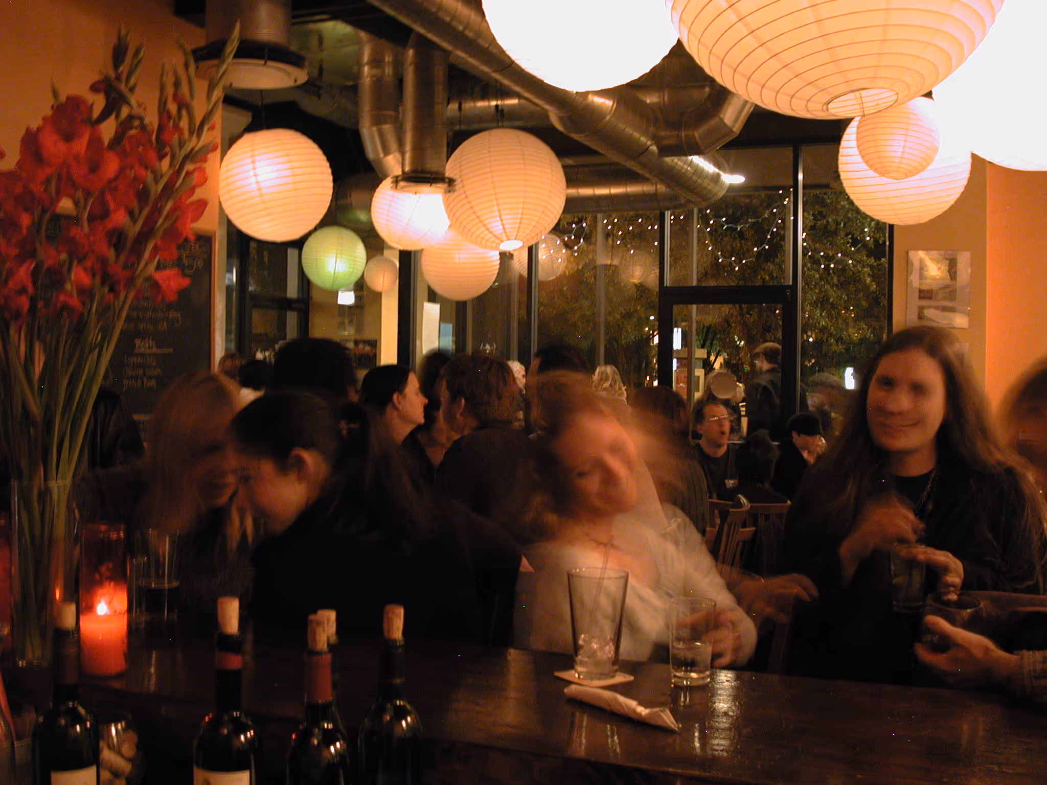 Dimly lit bar with people socializing, round paper lanterns hanging from the ceiling, and bottles of wine on the counter.
