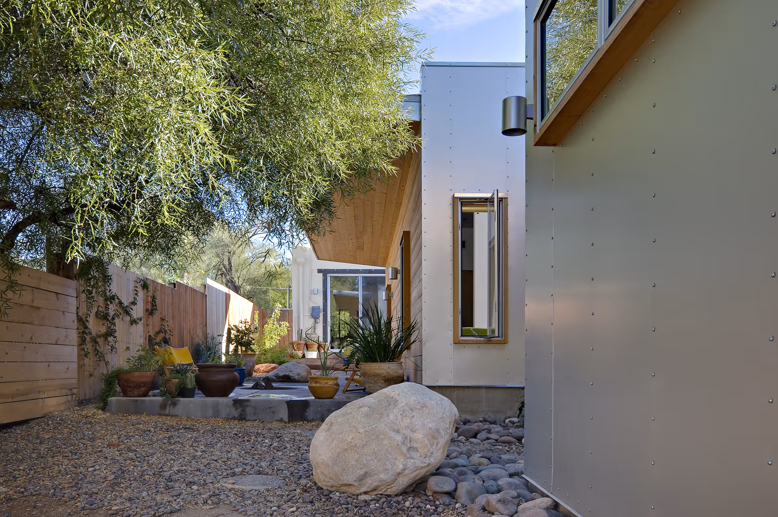 Modern backyard patio with potted plants, gravel ground, large rock, wooden fence, and open window on house.