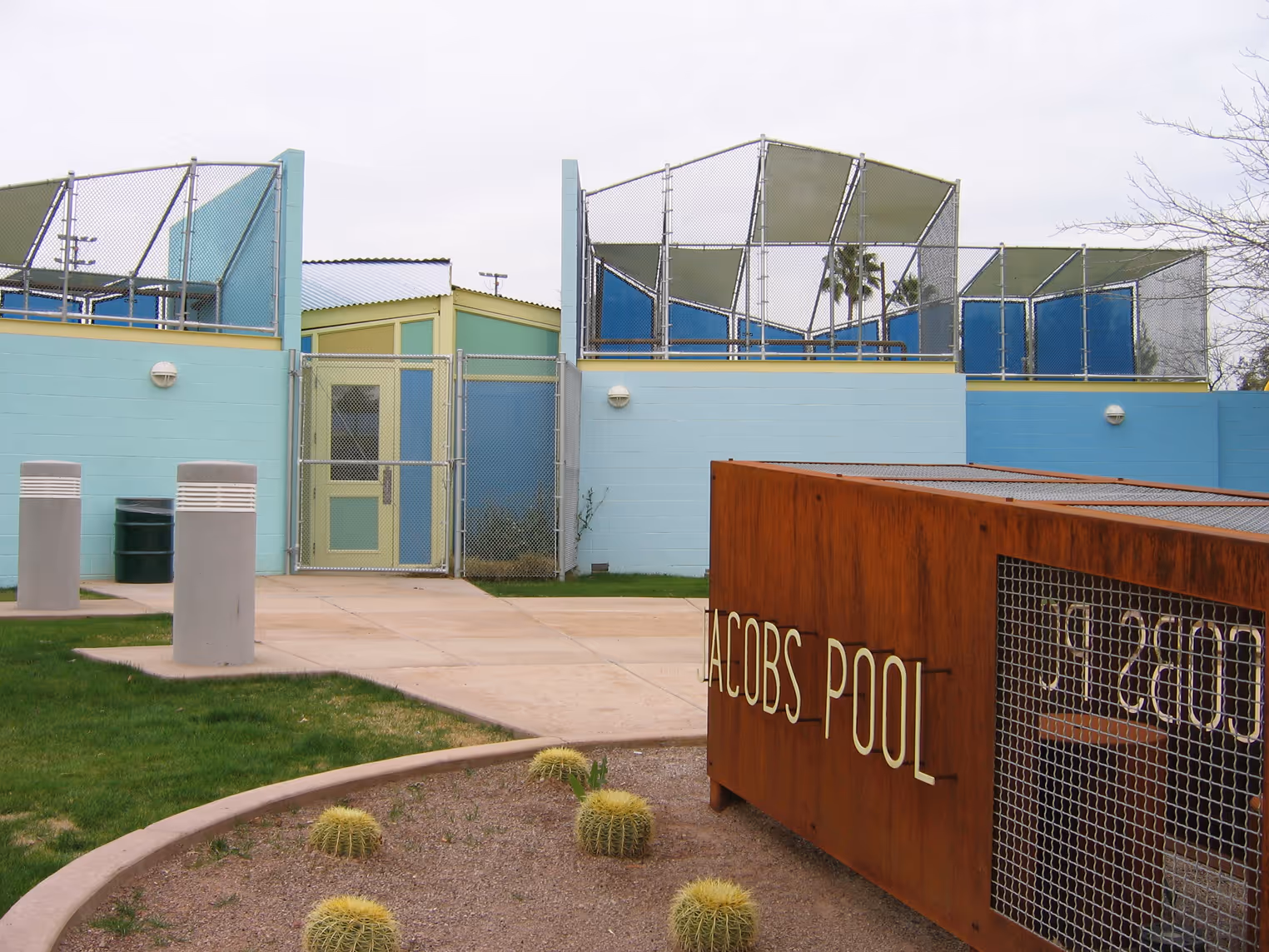 Outdoor view of a fenced area with light blue walls and a green door, with a rusty metal sign reading 'JACOBS POOL' near barrel cacti.