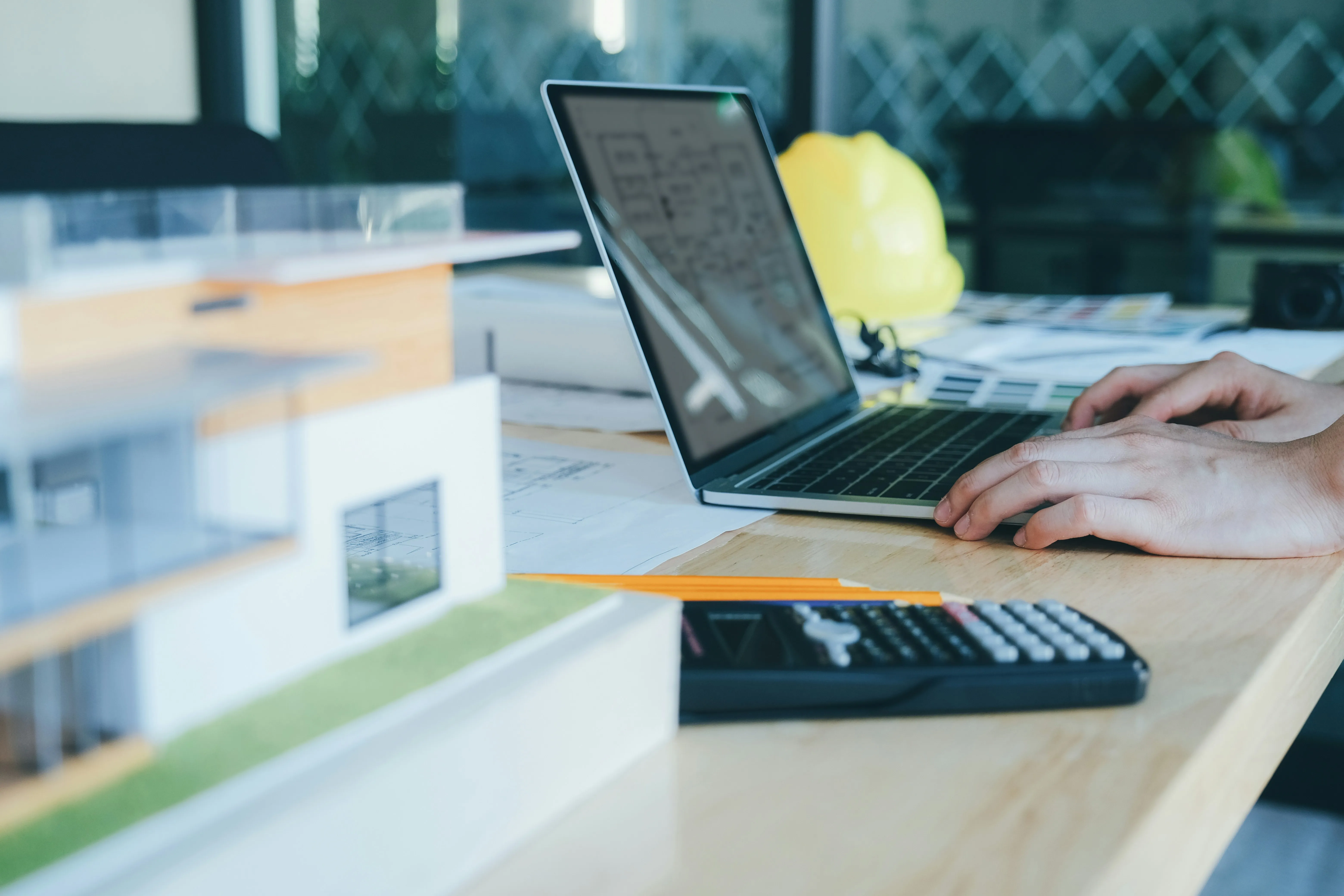 Person using laptop on a desk with architectural model, calculator, pencil, and yellow safety helmet in the background.