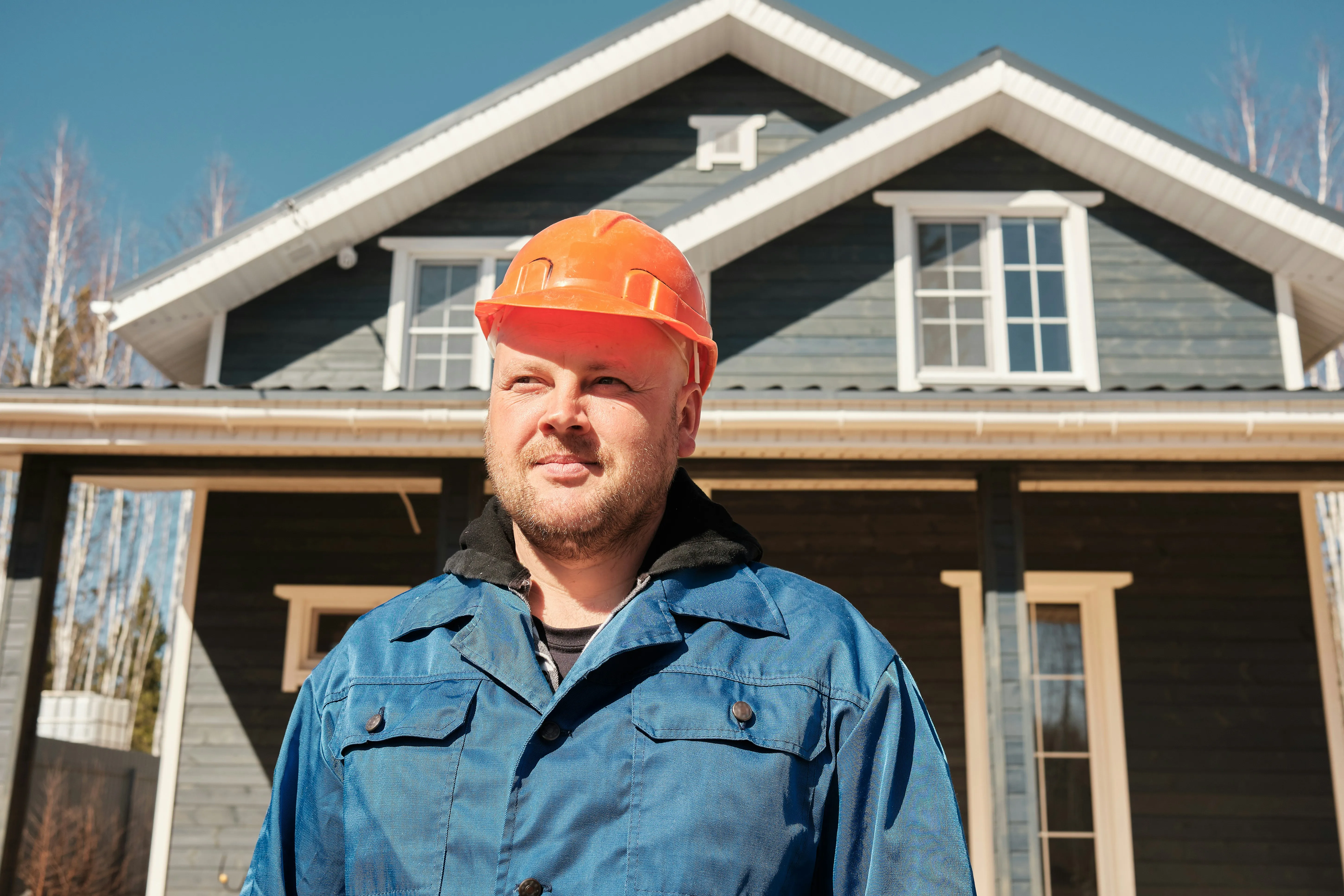 Construction worker in blue jacket and orange hard hat standing in front of a wooden house under clear blue sky.