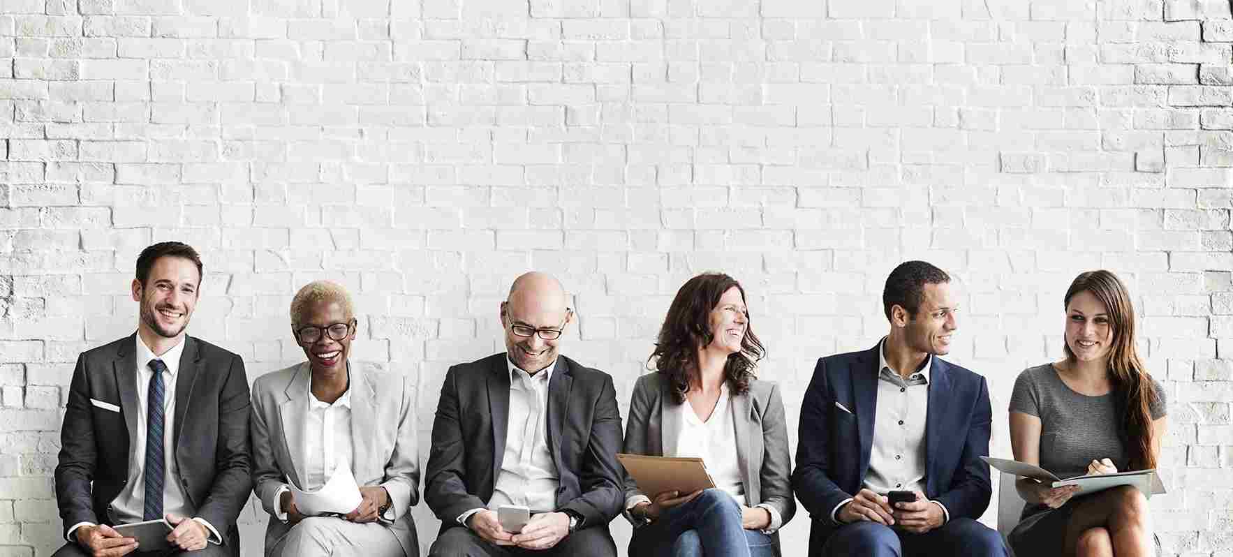 Sechs lächelnde Bewerberinnen und Bewerber in Businesskleidung sitzen vor einer weißen Ziegelwand