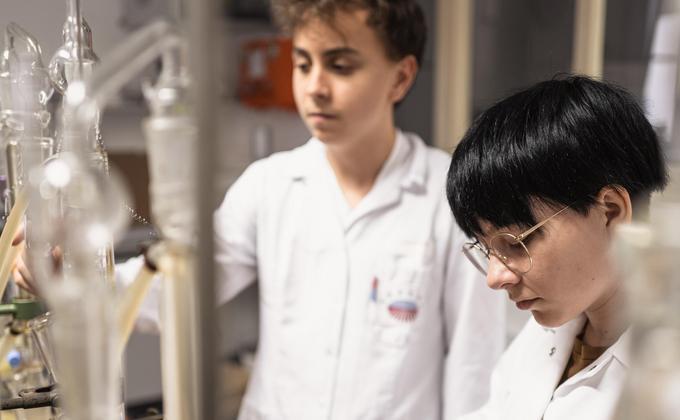 Two young lab technicians in white coats working behind glass apparatus in a laboratory