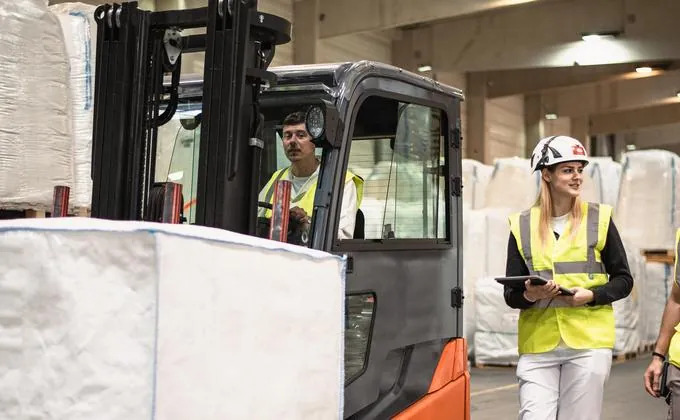 Forklift driver moving a big bag in a warehouse while a colleague in hard hat and safety vest checks a tablet