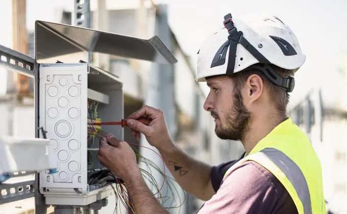 Electrician in a white hard hat and hi-vis vest wiring an outdoor switch cabinet