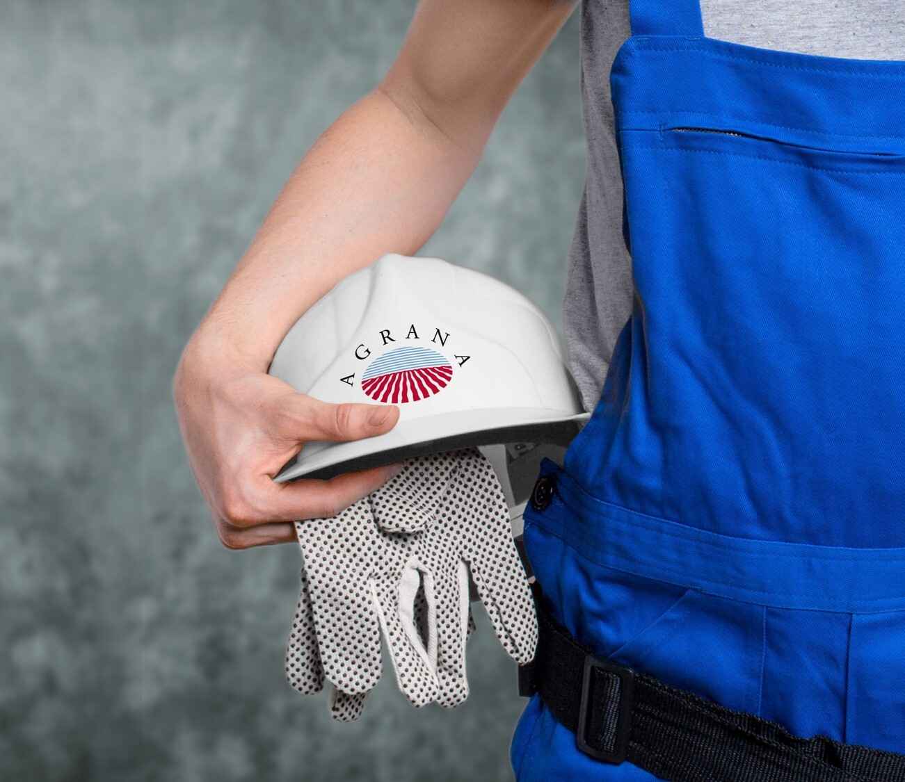 Worker in blue overalls holding a white AGRANA hard hat and protective gloves