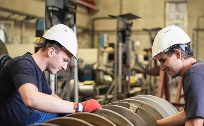 Two workers in white hard hats inspecting a large metal component in an industrial workshop