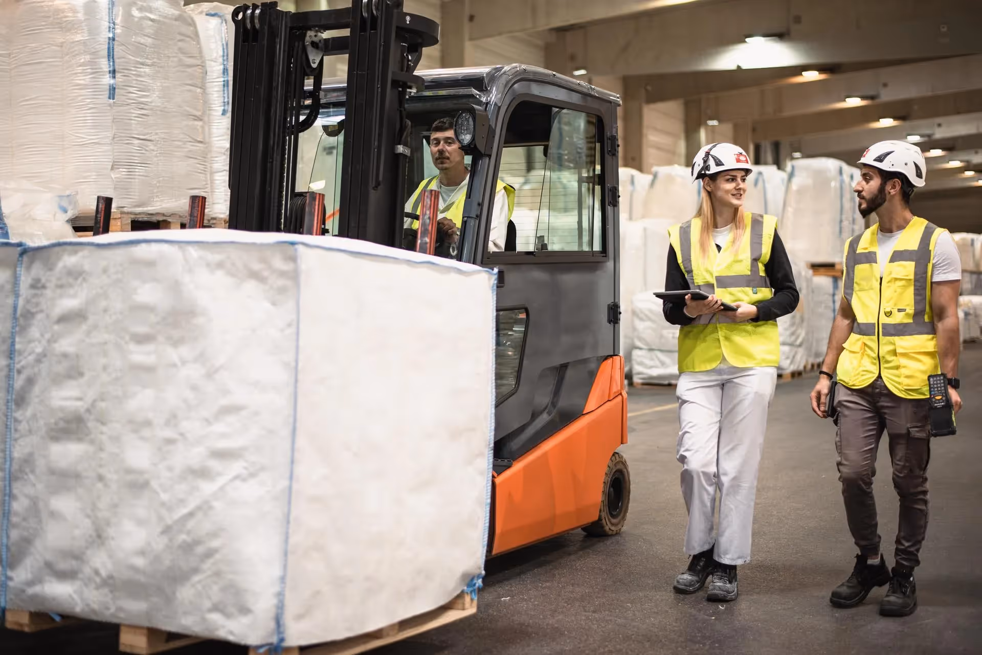Forklift moving big bags in a warehouse while two employees in hi-vis vests discuss a clipboard