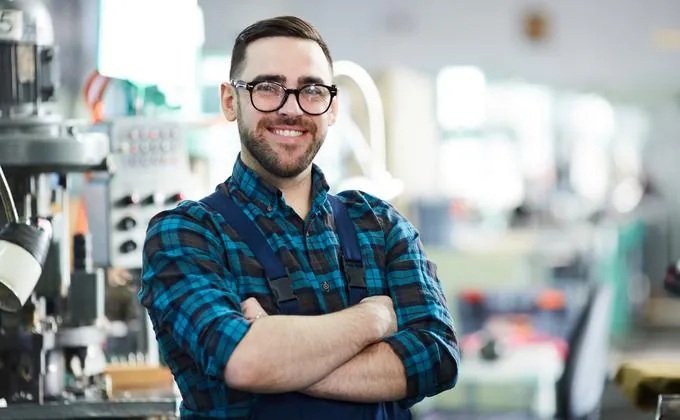 Lächelnder Techniker mit Brille und Karohemd, verschränkte Arme, in einer Produktionswerkstatt