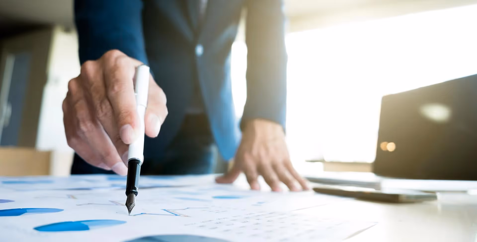 Businessman in a suit pointing a pen at charts on printed documents, laptop beside him