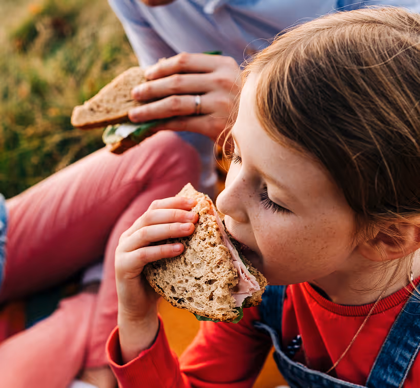 Girl in a red top biting into a wholegrain sandwich at a picnic in the grass