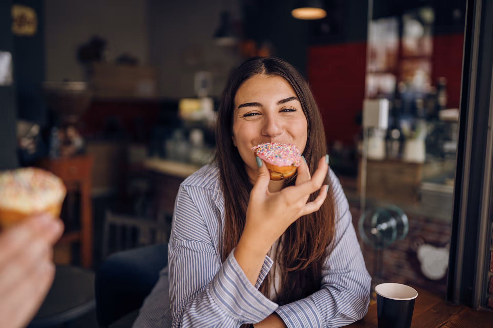 Lächelnde Frau beißt in einem Café in einen Donut mit rosa Streuseln
