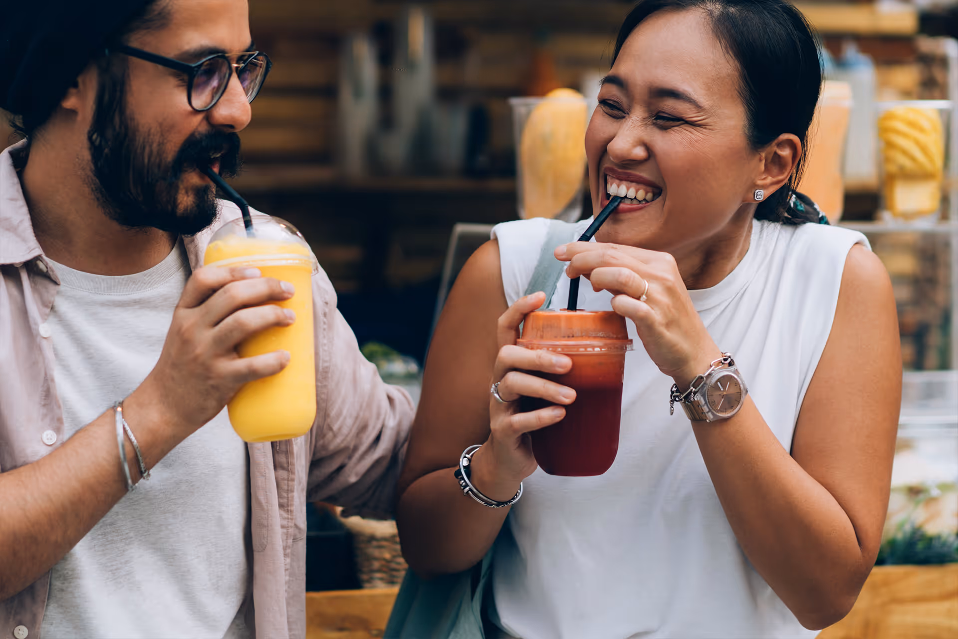 Lachendes Paar trinkt an einem Marktstand orangen und roten Fruchtsmoothie mit Strohhalm [no AI]