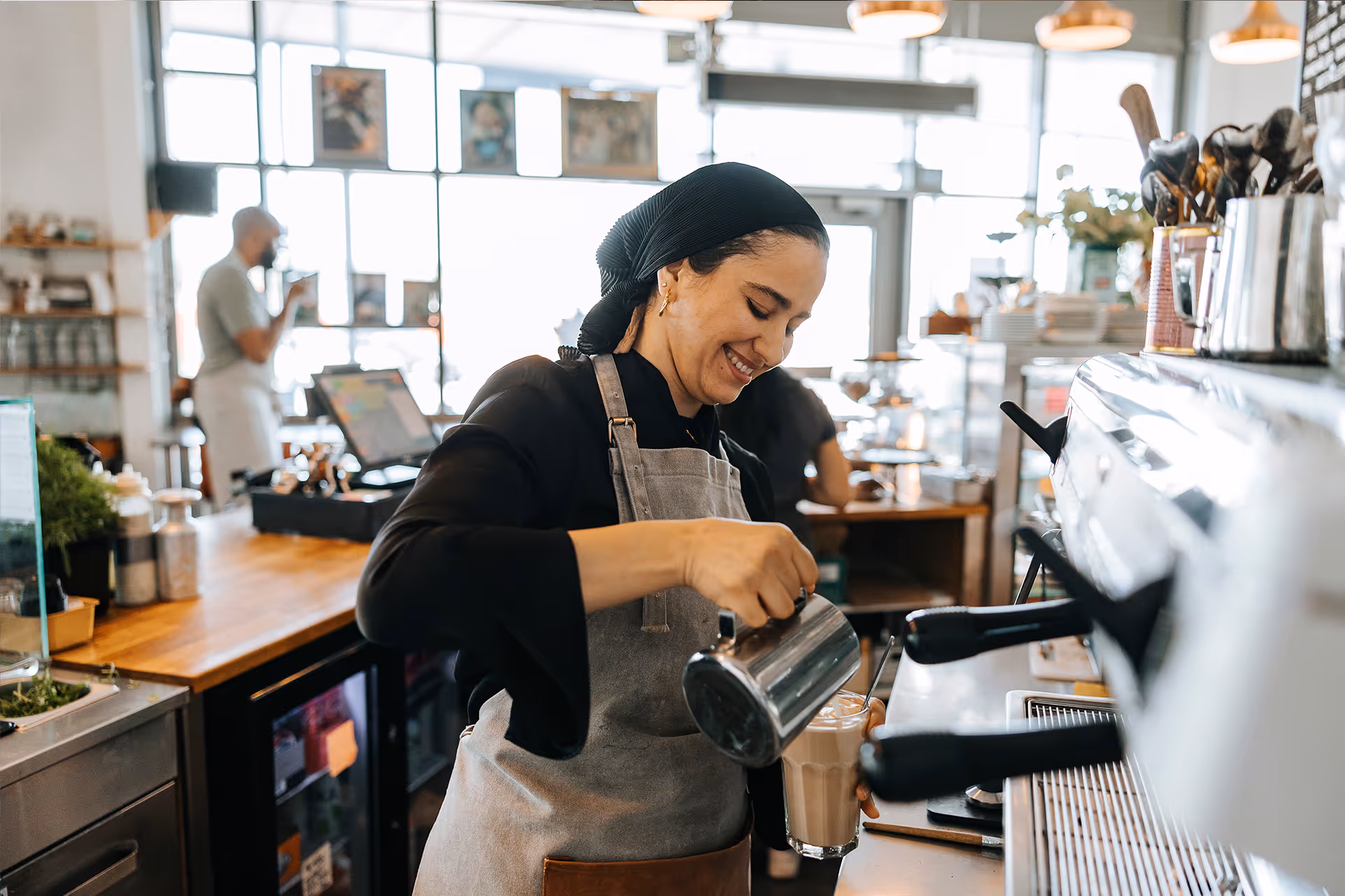 Lächelnde Barista mit Schürze gießt an der Espressomaschine Milch in einen Kaffee [no AI]