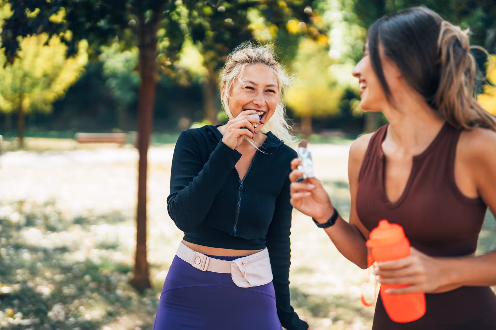 Zwei Frauen in Sportkleidung essen Energieriegel und halten eine Trinkflasche in einem sonnigen Park [no AI]