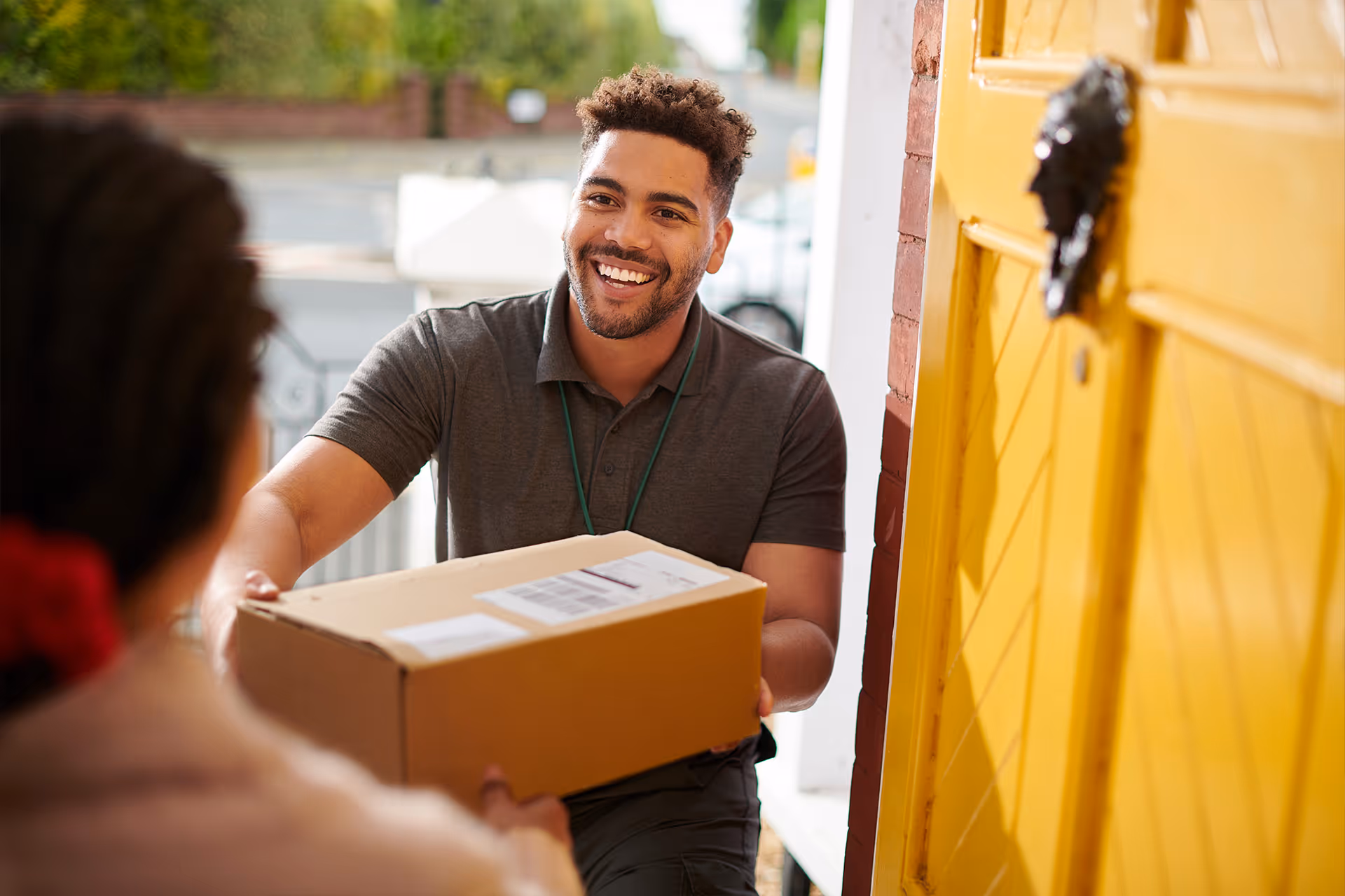 Lächelnder Paketbote übergibt ein Kartonpaket an einer gelben Haustür [no AI]