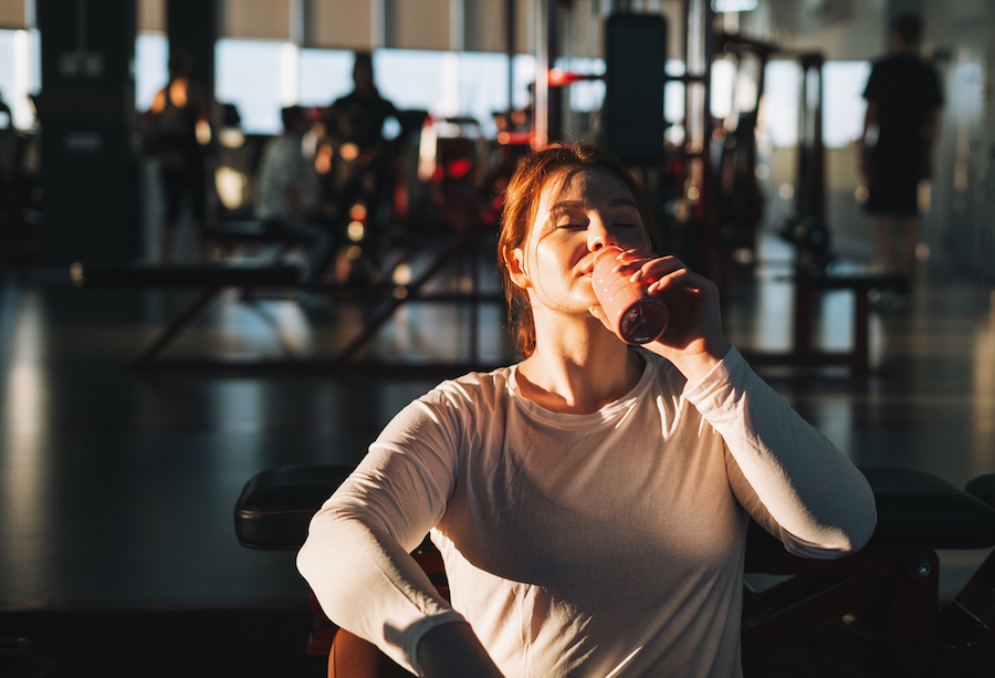 Frau trinkt in einem sonnendurchfluteten Fitnessstudio einen Proteinshake aus der Flasche [no AI]